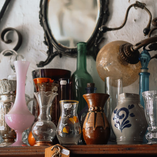 Collection of vintage vases and bottles on a wooden surface with a mirror and lamp in the background.