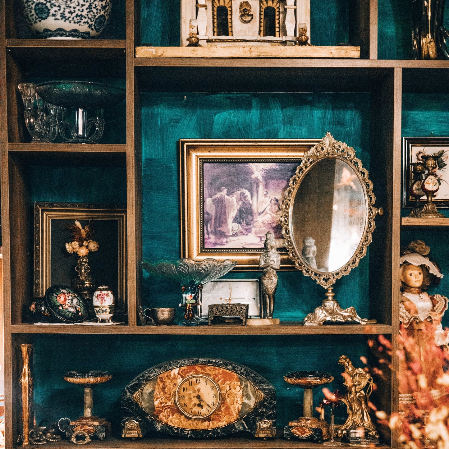a variety of home decor objects like small eggs, crystal bowls, vases, mirrors, clocks, and small picture frames arranged on a bookcase made of different shelf sizes and a greenish blue background
