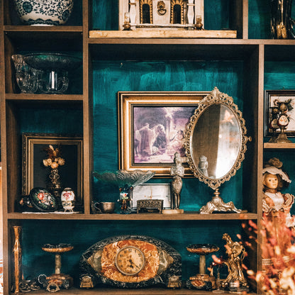 a variety of home decor objects like small eggs, crystal bowls, vases, mirrors, clocks, and small picture frames arranged on a bookcase made of different shelf sizes and a greenish blue background